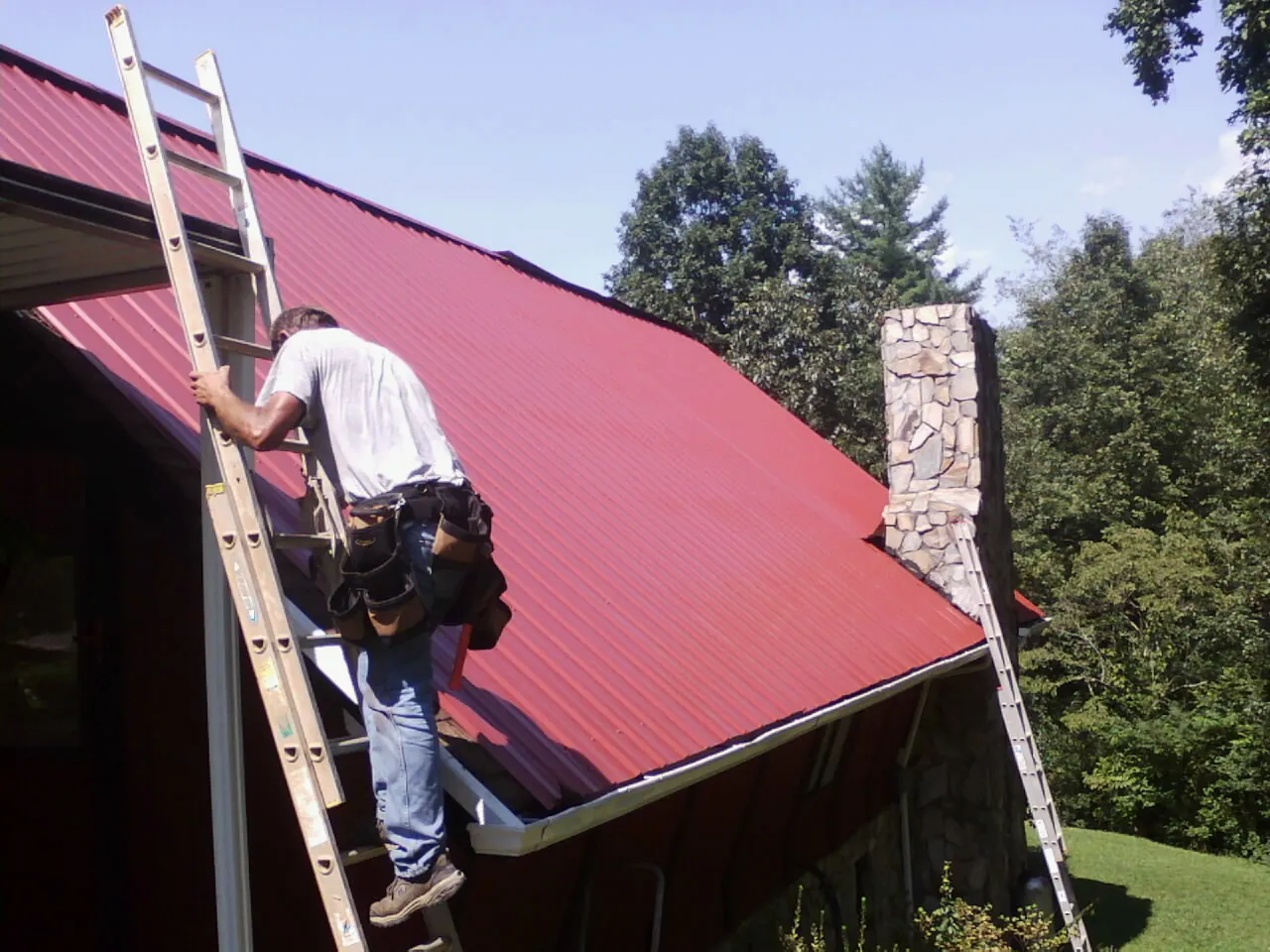 Roofer on a ladder beside a red metal roof and stone chimney — Soffit & Fascia Repair by Roofing Brothers in Mcville